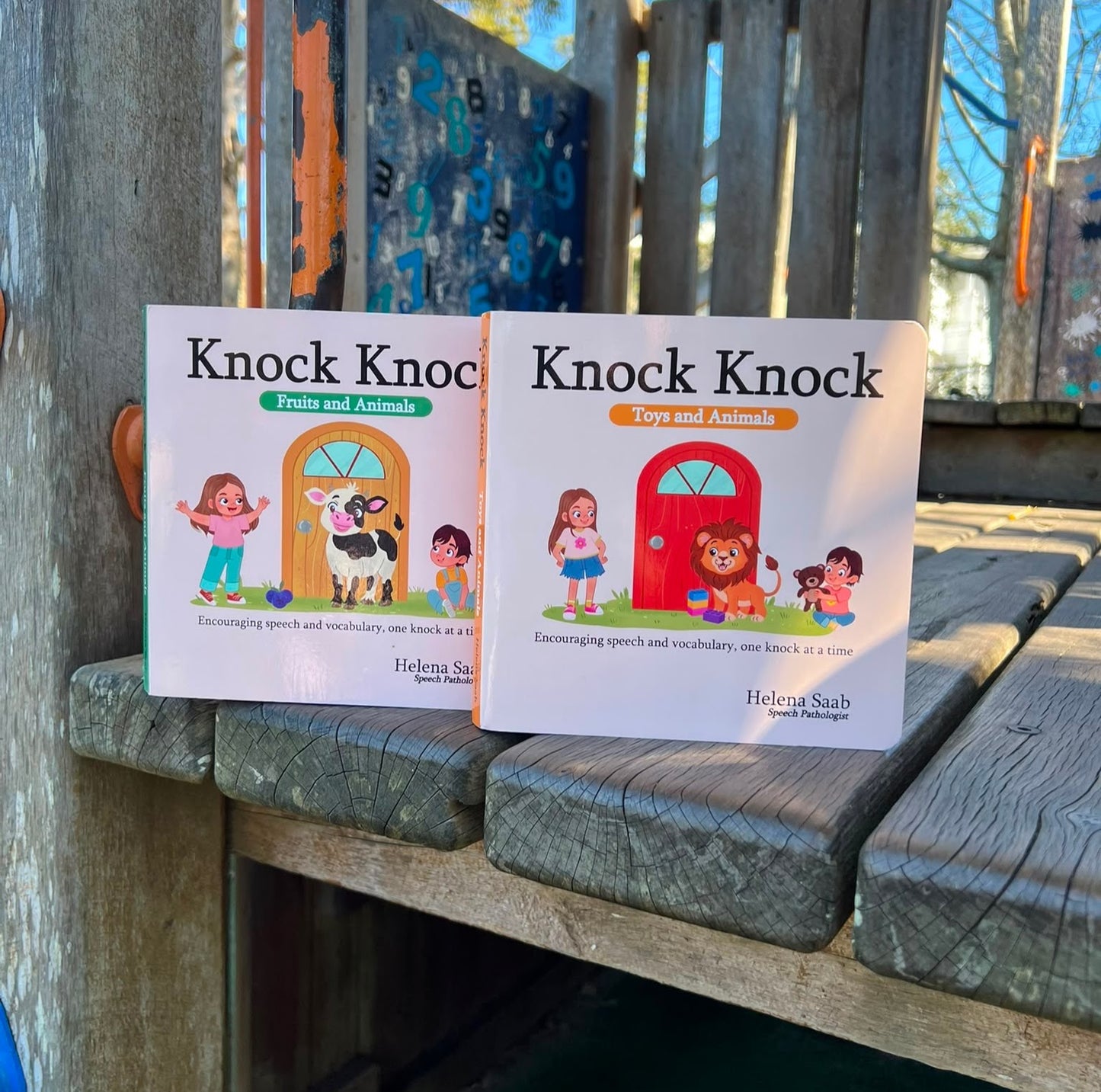 The Speechie Mumma Knock Knock board books, Fruits and Animals and Toys and Animals, displayed outdoors on a playground, encouraging speech and vocabulary development for toddlers