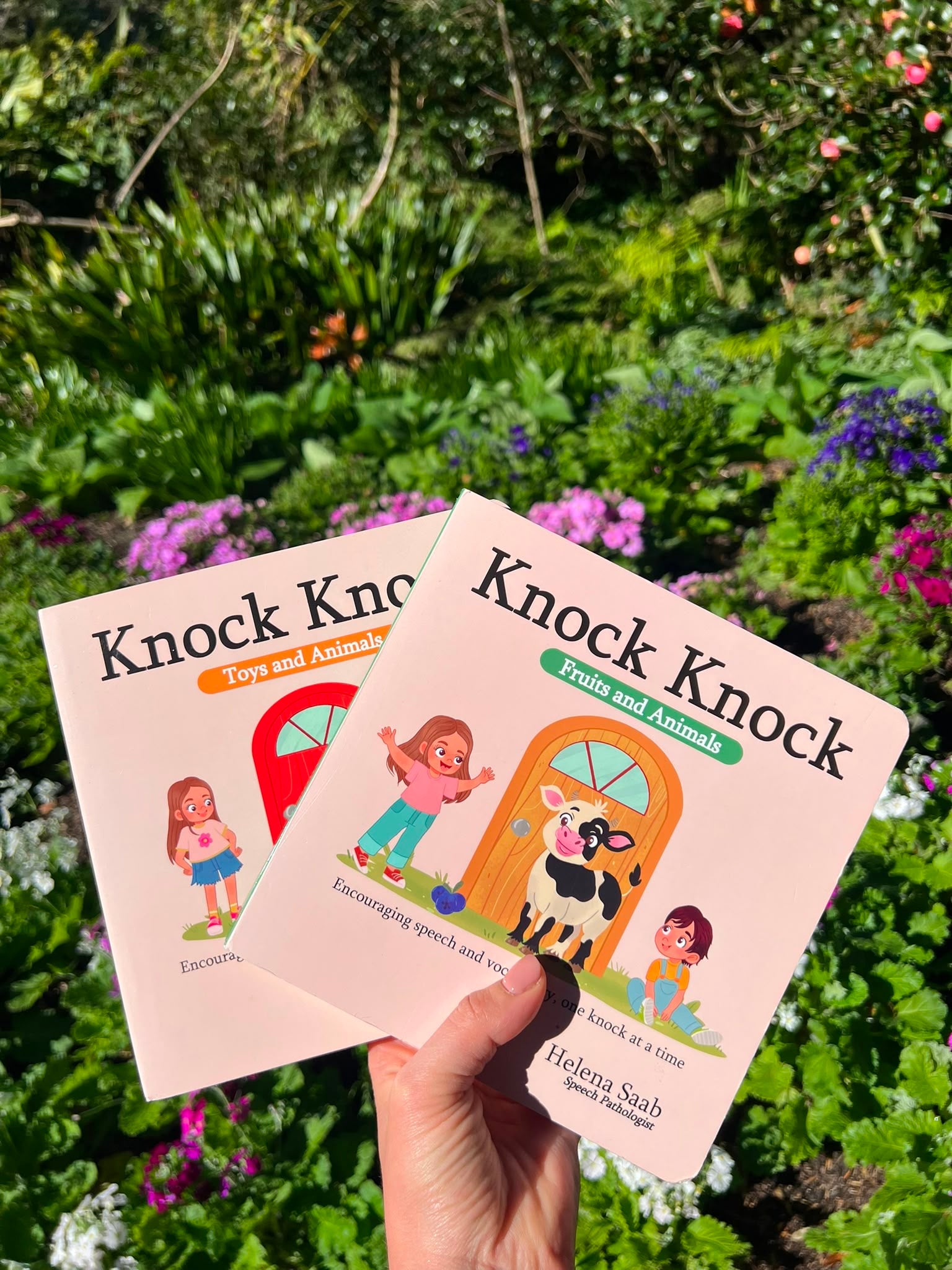 Hand holding the Speechie Mumma Knock Knock board books outdoors against blooming garden flowers, toddler speech development board books