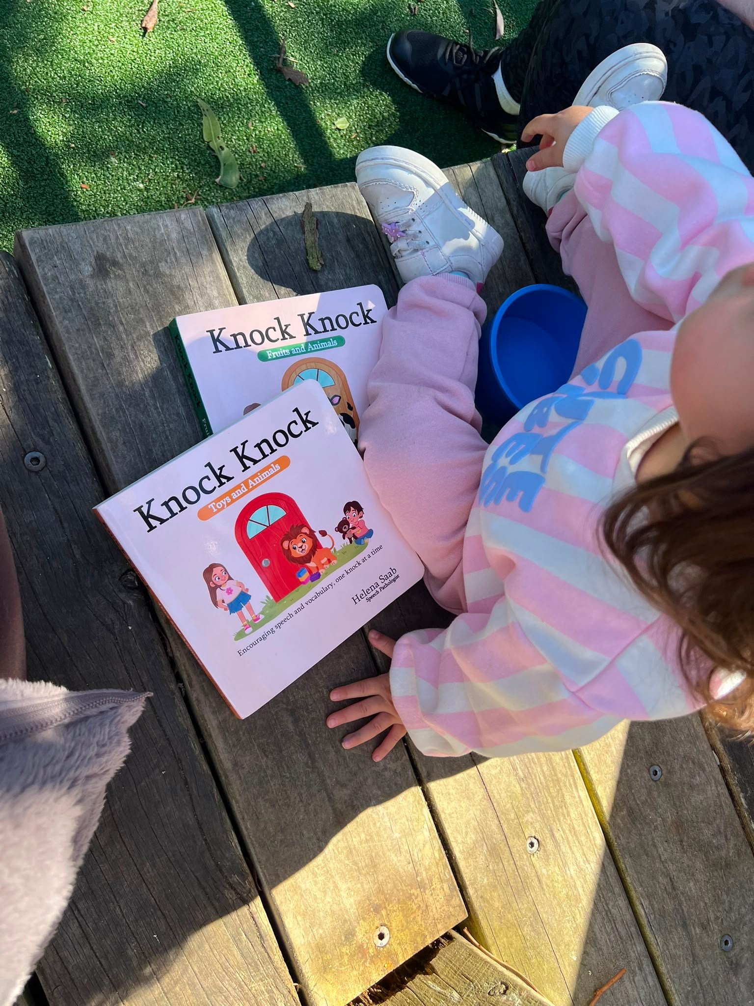 Kids sitting outdoors reading the Speechie Mumma Knock Knock board books, Fruits and Animals and Toys and Animals, everyday speech development for toddlers