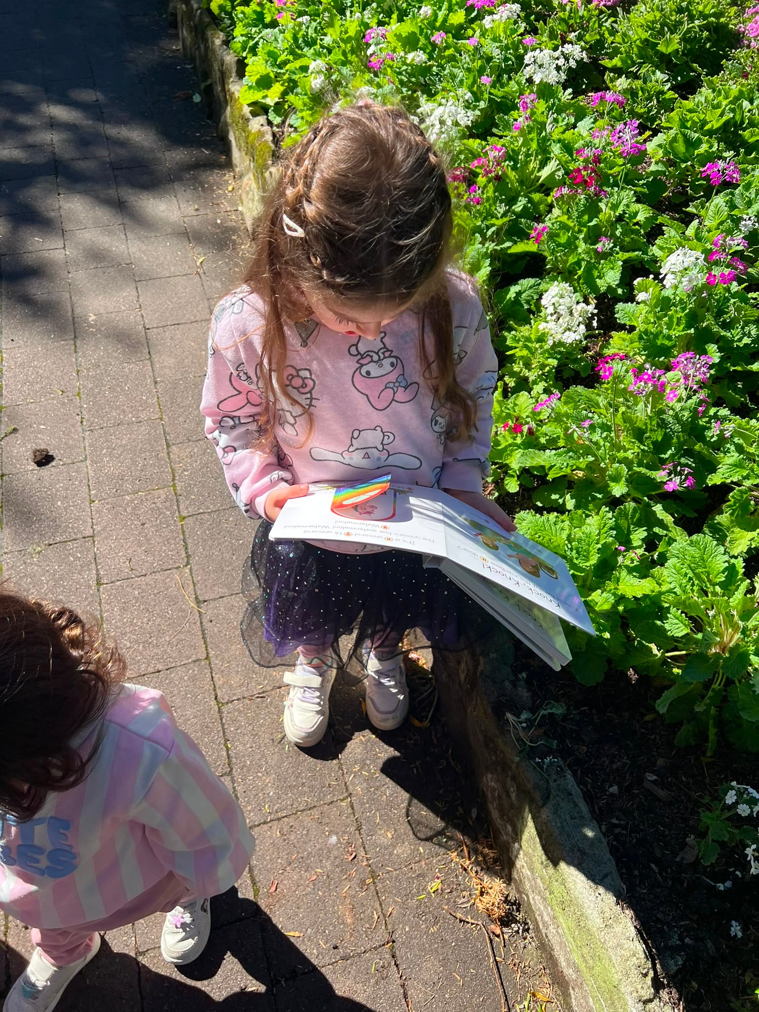 Toddler girl reading a Speechie Mumma Knock Knock board book outdoors among garden flowers, building early speech and vocabulary skills